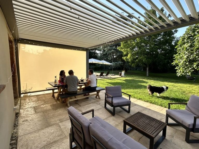 Personnes déjeunant à l'ombre sous la pergola adossée à lames orientables ouvertes et store latéral baissé